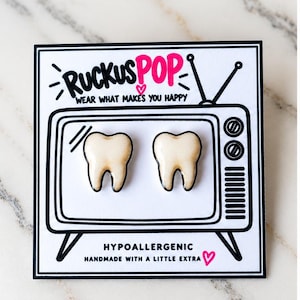 May include: A pair of off-white tooth-shaped stud earrings displayed on a white card featuring a black and white illustration of a vintage television set with the text RUCKUSPOP WEAR WHAT MAKES YOU HAPPY and HYPOALLERGENIC HANDMADE WITH A LITTLE EXTRA.