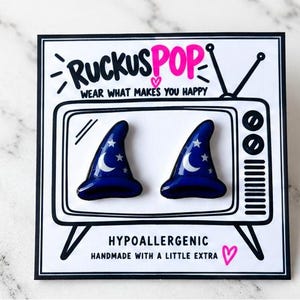Pode incluir: Um par de brincos em forma de chapéu de feiticeiro azul escuro com detalhes de lua crescente e estrelas brancas. Estão expostos num cartão branco com um gráfico de televisão, o texto RUCKUSPOP e WEAR WHAT MAKES YOU HAPPY.
