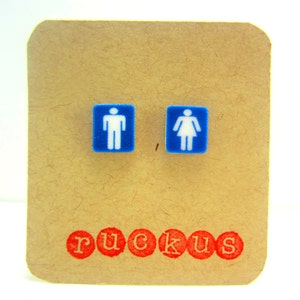 May include: A pair of blue and white earrings with a male and female restroom symbol design. The earrings are displayed on a brown card with the word "ruckus" printed in red.