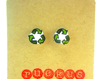 May include: A pair of small, white stud earrings with a green recycle symbol on each. The earrings are displayed on a brown card with the word "ruckus" printed in red.