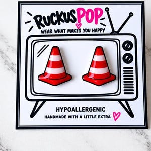 May include: A pair of red and white traffic cone stud earrings displayed on a white card featuring a black and white illustration of a vintage television set with the text RUCKUSPOP WEAR WHAT MAKES YOU HAPPY HYPOALLERGENIC HANDMADE WITH A LITTLE EXTRA.