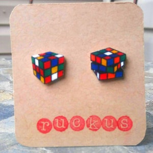 May include: A pair of colorful Rubik's Cube stud earrings displayed on a brown card. The earrings feature the classic cube design with red, blue, green, yellow, and white squares. The word "ruckus" is printed in red below the earrings.