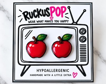 May include: A pair of red apple-shaped enamel pins with green leaves, displayed on a white backing card featuring a black line drawing of a vintage television set with the text RUCKUSPOP WEAR WHAT MAKES YOU HAPPY and HYPOALLERGENIC HANDMADE WITH A LITTLE EXTRA.