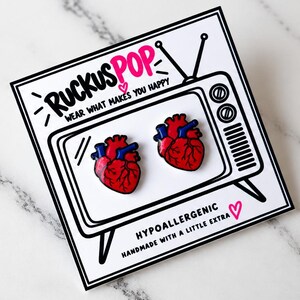 Pode incluir: Dois pins de esmalte em forma de cora&ccedil;&atilde;o anat&oacute;mico em vermelho e azul, apresentados num cart&atilde;o branco com um desenho a tra&ccedil;o preto de uma televis&atilde;o vintage e o texto RUCKUSPOP WEAR WHAT MAKES YOU HAPPY e HYPOALLERGENIC HANDMADE WITH A LITTLE EXTRA.