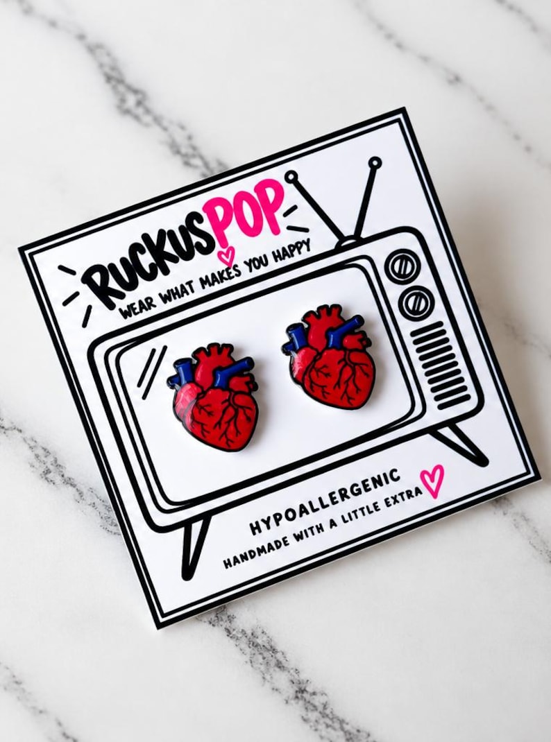 Pode incluir: Dois pins de esmalte em forma de cora&ccedil;&atilde;o anat&oacute;mico em vermelho e azul, apresentados num cart&atilde;o branco com um desenho a tra&ccedil;o preto de uma televis&atilde;o vintage e o texto RUCKUSPOP WEAR WHAT MAKES YOU HAPPY e HYPOALLERGENIC HANDMADE WITH A LITTLE EXTRA.