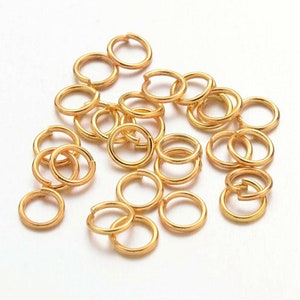 May include: A collection of small, gold-coloured jump rings, scattered on a white surface. The rings are open, ready for use in jewellery making or other crafts. The rings are round and have a shiny finish.