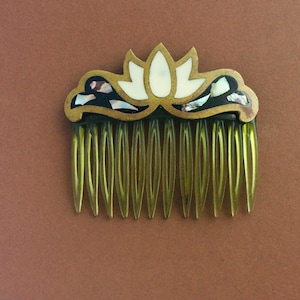 May include: A vintage hair comb with a decorative lotus flower design. The comb features a gold-toned frame with a black background, inlaid with iridescent shell pieces and a central white flower. The comb's teeth are a translucent amber color.
