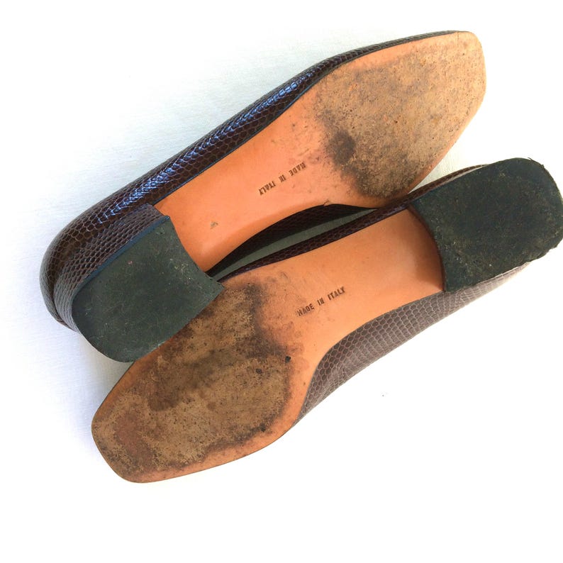 May include: A pair of brown, textured leather shoes with a low heel. The soles are a light brown color with the words "MADE IN ITALY" printed on them. The heels are dark green.