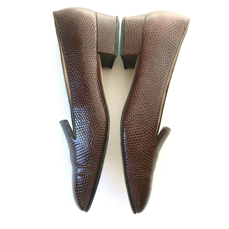 May include: A pair of brown leather loafers with a textured, reptile-like pattern. The shoes have a low heel and a classic slip-on design, perfect for everyday wear. The shoes are displayed on a white background.