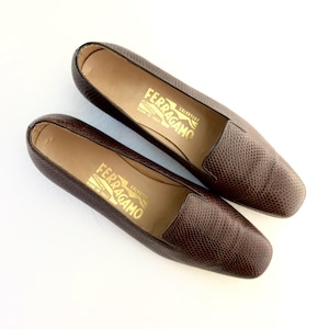 May include: A pair of brown leather loafers with a textured, snakeskin-like pattern. The shoes have a square toe and a low heel. The brand name "FERRAGAMO" is printed in gold on the insole.