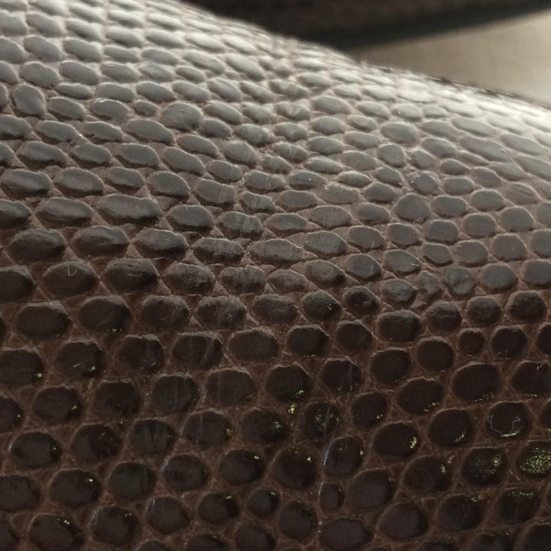 May include: Close-up of a dark brown, textured surface with a repeating pattern of small, rounded shapes. The surface has a slight sheen, suggesting a leather or similar material. The pattern resembles reptile skin.