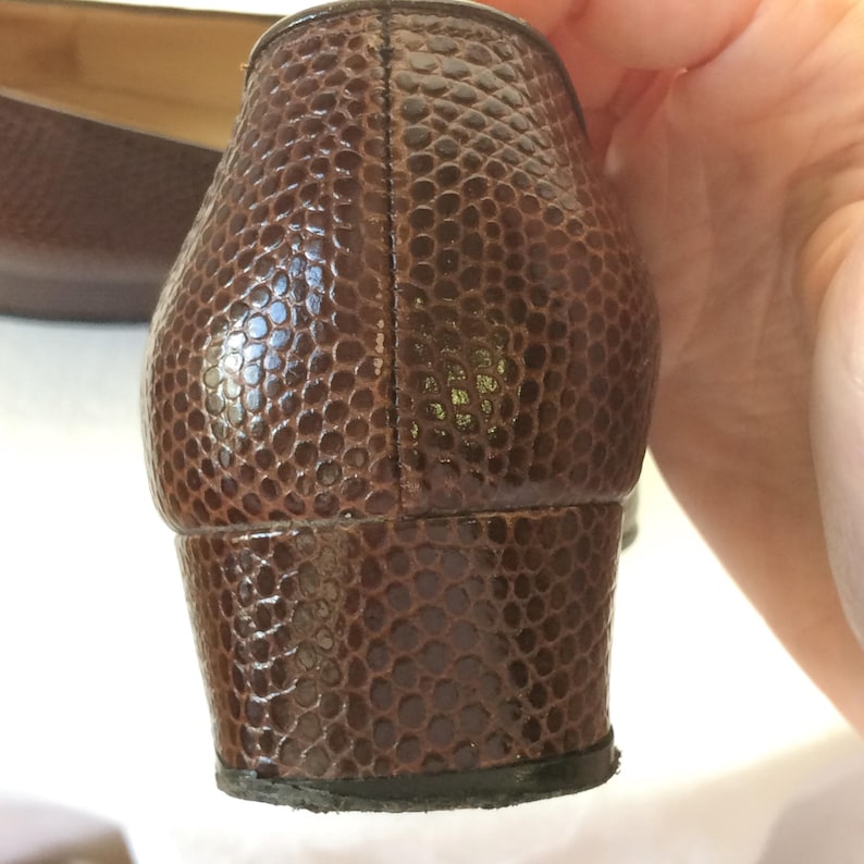 May include: Close-up of a brown, textured shoe heel. The shoe has a reptile-like pattern and a small, block heel. The shoe appears to be leather and is a classic style.