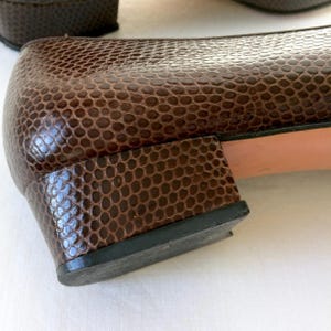 May include: Close-up of a pair of brown leather shoes with a reptile-like texture. The shoes have a low heel and a black sole. The leather has a glossy finish.