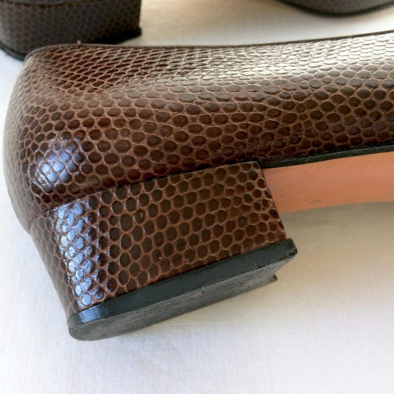 May include: Close-up of a pair of brown leather shoes with a reptile-like texture. The shoes have a low heel and a black sole. The leather has a glossy finish.