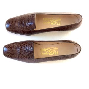 May include: A pair of brown leather loafers with a textured, reptile-like pattern. The shoes have a rounded toe and a low heel. The brand name "FERRAGAMO" is printed in gold on the insole.