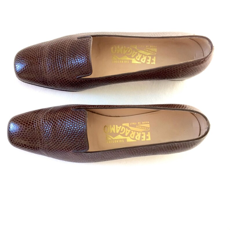May include: A pair of brown leather loafers with a textured, reptile-like pattern. The shoes have a rounded toe and a low heel. The brand name "FERRAGAMO" is printed in gold on the insole.