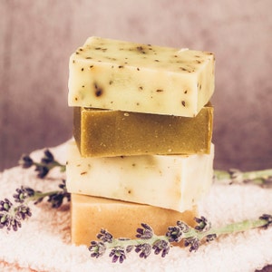 May include: Four bars of natural soap stacked on top of each other. The top bar is green and white, the second bar is green, the third bar is white, and the bottom bar is beige. The bars of soap are sitting on a white towel with lavender flowers.