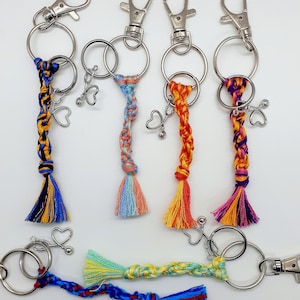 May include: Five colourful braided keychains with silver key rings and charms. Each keychain has a tassel at the end. The colours include blue, yellow, orange, red, and purple.