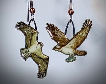 May include: A pair of silver earrings featuring a detailed image of an osprey with a fish in its talons. The osprey is depicted in flight with its wings spread wide. The earrings are suspended from silver hooks with a copper bead at the top.