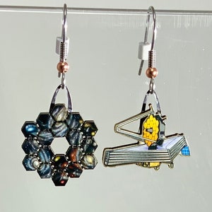 May include: A pair of silver earrings with a space-themed design. One earring features a hexagonal design with images of galaxies and nebulae. The other earring features a silver model of the Hubble Space Telescope.