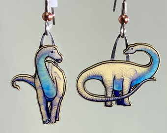 May include: A pair of silver earrings featuring a blue and white dinosaur design. The dinosaurs are depicted in a cartoon style with long necks and tails.