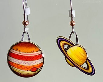 May include: A pair of silver earrings with a colorful depiction of Jupiter and Saturn. The Jupiter earring is orange, red, and white with a swirling pattern. The Saturn earring is yellow with a purple ring.