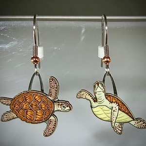 May include: A pair of silver dangle earrings featuring a detailed illustration of a sea turtle. The turtle is painted in shades of brown, yellow, and white. The earrings are suspended from a silver hook with a small copper bead.