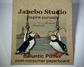 May include: A pair of dangle earrings featuring a colorful illustration of Atlantic Puffins. The earrings are attached to a brown post-consumer paperboard card with the text "Jabebo Studio" and "inspire curiosity". The puffins have black, white, and orange details.