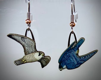 May include: A pair of silver earrings with a blue and white bird design. The birds are in flight and have detailed wings and beaks.