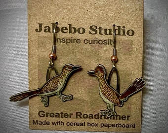May include: A pair of earrings featuring a roadrunner design. The earrings are made from cereal box paperboard and are labeled "Greater Roadrunner" and "Jabebo Studio inspire curiosity".