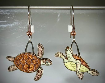 May include: A pair of silver dangle earrings featuring a detailed illustration of a sea turtle. The turtle is painted in shades of brown, yellow, and white. The earrings are suspended from a silver hook with a small copper bead.