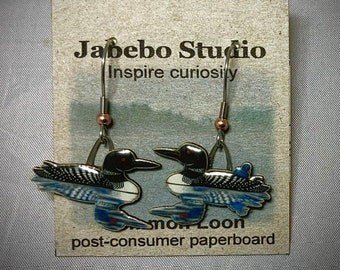 Puede incluir: Pendientes colgantes con diseño de colimbo. Los pendientes tienen detalles de esmalte negro, blanco y azul. Cuelgan de ganchos plateados con una cuenta de color cobre. Los pendientes se exhiben en un respaldo de cartón con el texto "Jabebo Studio" e "Inspire curiosity".