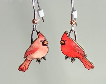 May include: A pair of dangle earrings featuring red cardinal birds with silver hooks and copper beads. The birds are painted in a realistic style with detailed feathers and beaks.