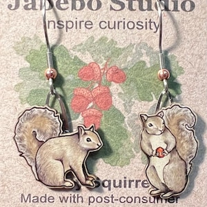 May include: Squirrel earrings with a whimsical design. Each earring features a detailed illustration of a squirrel holding a nut. The earrings are made from post-consumer cereal box paperboard and have silver-colored hooks.