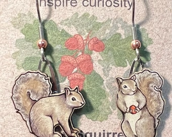May include: Squirrel earrings with a whimsical design. Each earring features a detailed illustration of a squirrel holding a nut. The earrings are made from post-consumer cereal box paperboard and have silver-colored hooks.