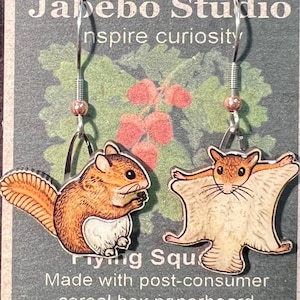 May include: A pair of earrings featuring a flying squirrel design. The earrings are made from recycled cereal box paperboard and have silver hooks. The text "Flying Squirrels" and "Made with post-consumer cereal box paperboard" is printed on the card.