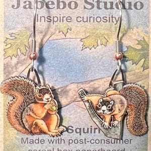 May include: A pair of earrings featuring a squirrel design made from recycled cardboard. The earrings are silver with a brown and black squirrel design. The text "Squirrel" and "Made with post-consumer cereal box paperboard" is printed on the card.