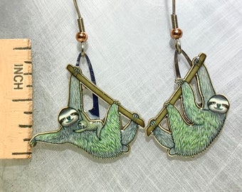 May include: A pair of silver earrings featuring a green sloth with a baby sloth hanging on its back. The sloths are depicted hanging from a branch.