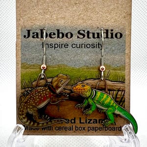 May include: A pair of earrings featuring a green and yellow lizard and a brown and red lizard. The earrings are made with cereal box paperboard and are hanging from silver hooks. The card reads "Jabebo Studio inspire curiosity" and "Painted Lizard Made with cereal box paperboard".