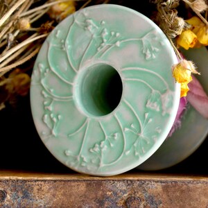 May include: A green ceramic spool with a floral design. The spool has a hole in the centre and is sitting on a wooden surface.