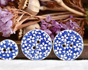 May include: Three ceramic buttons with a blue and white floral pattern. The buttons are round with two holes and vary in size. Dried purple flowers and seed pods are in the background.