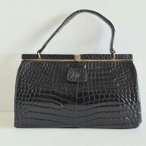 May include: A black handbag with a glossy, textured crocodile-skin pattern. The bag features a single top handle, a rectangular frame, and a gold-toned metal clasp closure centered on the top edge. The surface has a distinct, repeating scale-like texture.