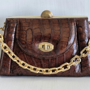 May include: A brown handbag with a textured crocodile-embossed pattern, featuring a gold-tone metal chain strap, a central gold-tone twist-lock closure on the front flap, and a semi-circular gold-tone clasp on the top frame.