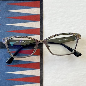 May include: Eyeglasses with a rectangular frame and a patterned, iridescent finish. The temples are dark, and the lenses are clear. The glasses rest on a blue, red, and white patterned surface. The text "OGI EVOLUTION" is visible.