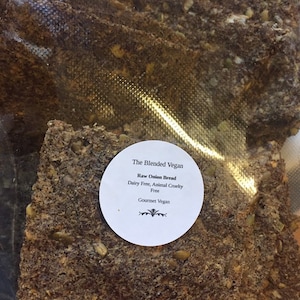 Op de afbeelding: Close-up van een verpakking rauw uienbrood. Het brood is donkerbruin met zichtbare zaden en granen. Een wit rond label leest "The Blended Vegan" en vermeldt "Raw Onion Bread, Dairy Free, Animal Cruelty Free, Gourmet Vegan."
