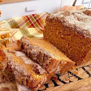 May include: A loaf of pumpkin bread, partially sliced, dusted with sugar. The bread sits on a wooden cutting board with the word "GRATE" visible. A plaid cloth and a pumpkin are in the background.