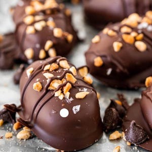 May include: Close-up of chocolate-covered peanut butter cups with chopped peanuts and a drizzle of chocolate on top. The cups are arranged on a silver surface.