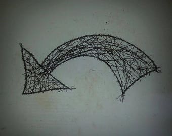May include: A black metal arrow shape made of wire, with a curved top and a pointed tip. The arrow is attached to a white wall.
