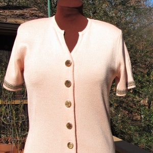 May include: A pale pink short-sleeved cardigan with a brown and white geometric pattern at the hem. The cardigan has a button-up front with gold buttons.