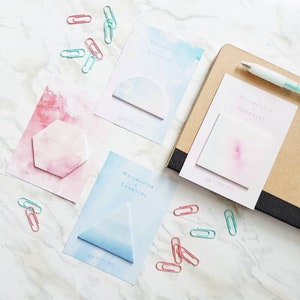 Puede incluir: Surtido de notas adhesivas de colores con formas geométricas y diseños de acuarela. Las notas incluyen el texto "Watercolor & Geometry" y el nombre de la marca "Sticky Note Cafe".